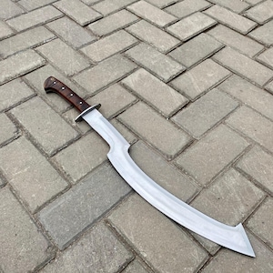 May include: A curved, silver-colored sword with a brown wooden handle. The sword is lying on a gray brick surface.