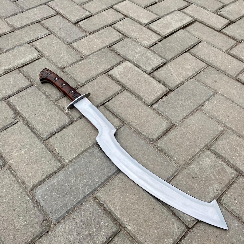 May include: A curved, silver-colored sword with a brown wooden handle. The sword is lying on a gray brick surface.