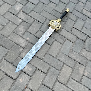 Puede incluir: Una espada decorativa con una hoja plateada y una empu&ntilde;adura dorada. La empu&ntilde;adura presenta un dise&ntilde;o ornamentado con una piedra transparente central. El mango est&aacute; envuelto en material negro. La espada est&aacute; sobre una superficie de ladrillos grises.