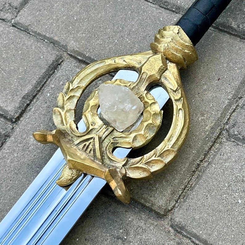 Puede incluir: Una espada decorativa con una hoja de acero pulido y una empu&ntilde;adura dorada. La empu&ntilde;adura presenta un dise&ntilde;o ornamentado con un cristal central de color claro. El mango est&aacute; envuelto en material negro.