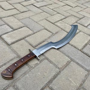 May include: A curved, silver-colored sword with a wooden handle. The handle is brown with a dark wood grain pattern. The sword is lying on a gray brick surface.