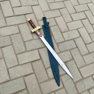 Op de afbeelding: Een zilveren zwaard met een bruin en gouden handvat en een rood embleem op het lemmet, naast een zwarte leren schede. Het zwaard heeft een decoratief ontwerp op het lemmet. De schede is van leer.