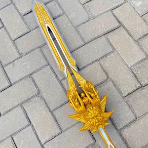 May include: A decorative sword with a silver blade and gold-colored hilt. The hilt features an ornate design with a detailed figure and a starburst shape. The sword is lying on a gray brick surface.