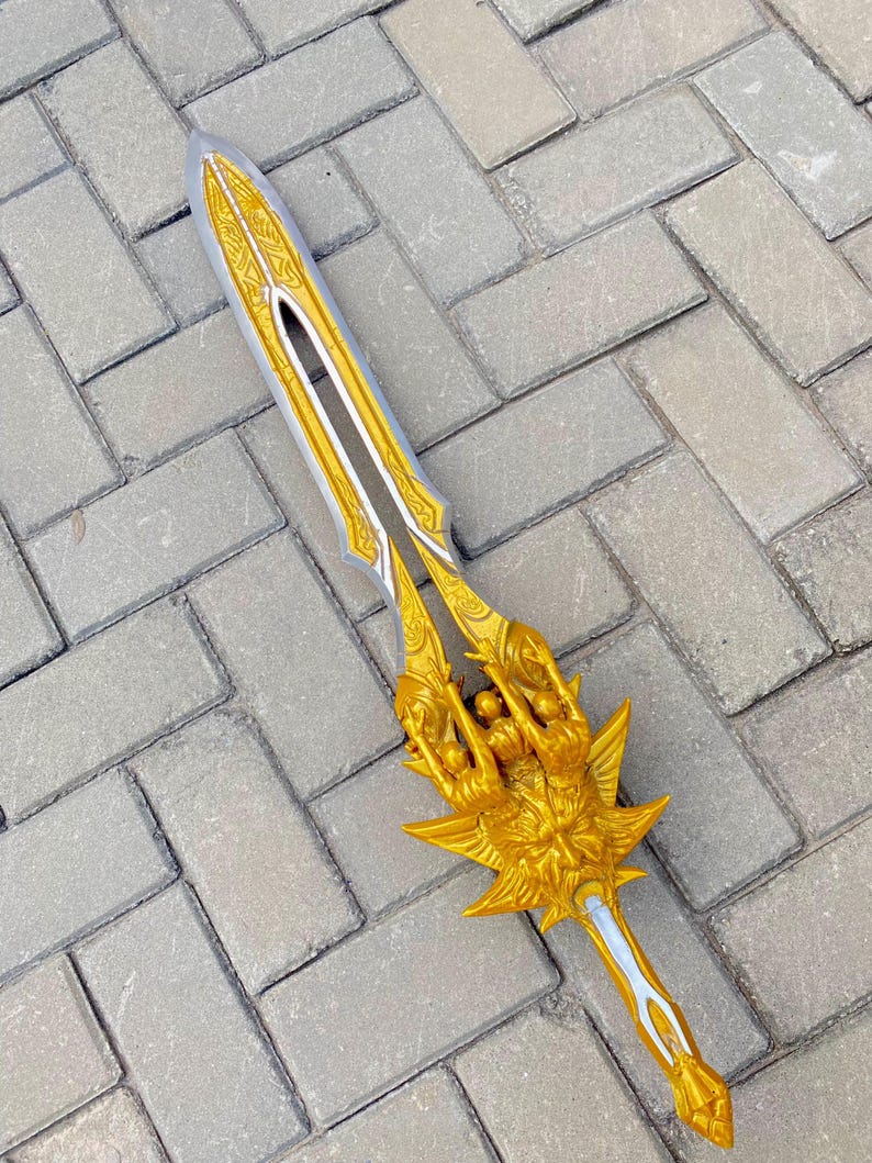May include: A decorative sword with a silver blade and gold-colored hilt. The hilt features an ornate design with a detailed figure and a starburst shape. The sword is lying on a gray brick surface.