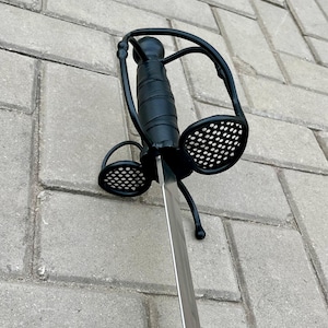 Könnte beinhalten: Ein schwarzes Rapier-Schwert mit aufwendigem Parier. Der Griff ist mit schwarzem Material umwickelt, und der Parier hat zwei kreisförmige Lochplatten. Die Klinge ist silberfarben und zeigt nach unten.