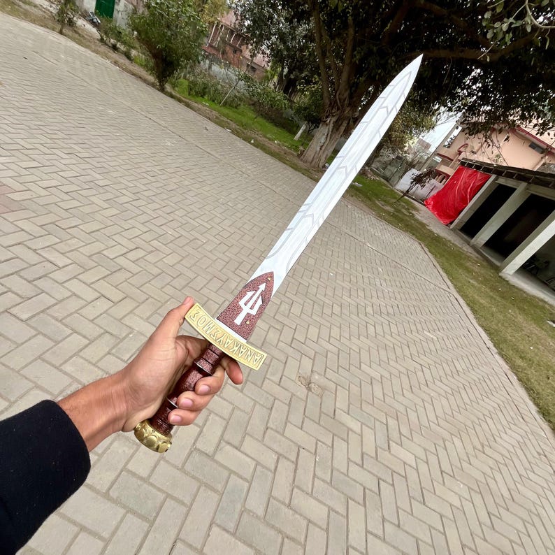 Puede incluir: Una espada plateada y dorada con un mango marr&oacute;n se sostiene en una mano. La espada presenta un s&iacute;mbolo de tridente en la guarda y un mango decorativo. La hoja mide aproximadamente 76 cm de largo.