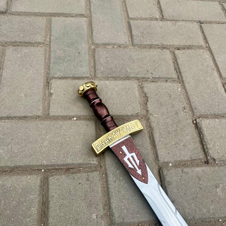 Puede incluir: Una espada decorativa con empu&ntilde;adura marr&oacute;n y guarda dorada. La hoja es plateada con un s&iacute;mbolo de tridente y la palabra griega "ANAKAY&Sigma;MO&Sigma;" en la guarda. La espada est&aacute; sobre una superficie de ladrillos grises.