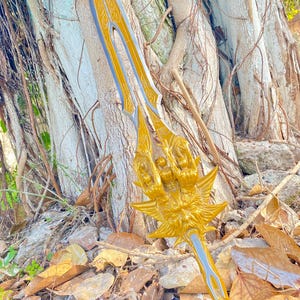 May include: A golden fantasy sword with intricate details, featuring a stylized handle and blade design. The sword has a gold finish with silver accents and is set against a natural background of a tree and fallen leaves.