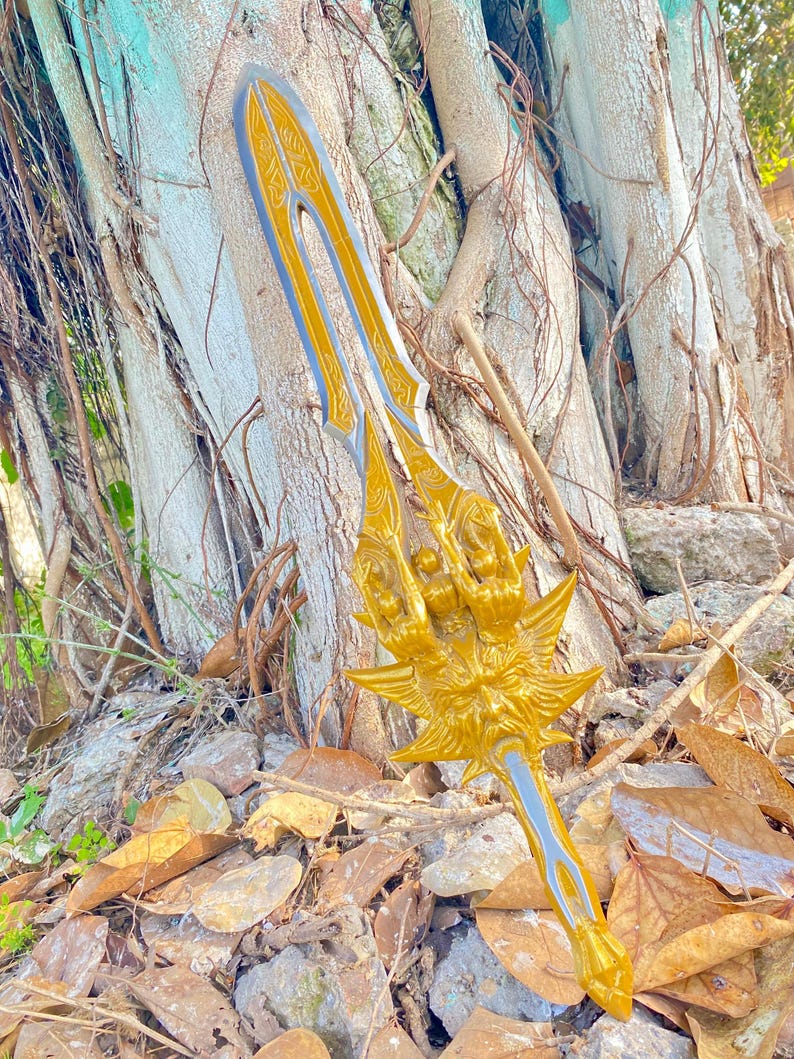 May include: A golden fantasy sword with intricate details, featuring a stylized handle and blade design. The sword has a gold finish with silver accents and is set against a natural background of a tree and fallen leaves.