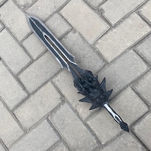 May include: A black and silver fantasy sword with intricate details. The blade has a central groove and silver accents. The hilt features a detailed dragon design. The sword is lying on a gray brick surface.