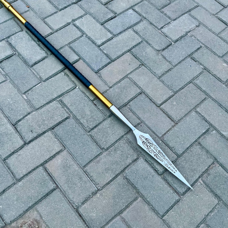May include: A spear with a silver blade and a black and gold shaft. The blade has an ornate design etched into it. The shaft is black with gold accents near the blade and at the top. The spear is lying on a gray brick surface.