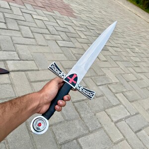 May include: A silver and black sword with a pointed blade. The hilt is black with a red cross design. The crossguard is silver with a decorative pattern. The pommel is silver with a red gem. The sword is held in a hand.
