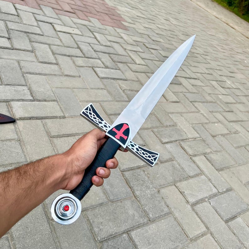 May include: A silver and black sword with a pointed blade. The hilt is black with a red cross design. The crossguard is silver with a decorative pattern. The pommel is silver with a red gem. The sword is held in a hand.