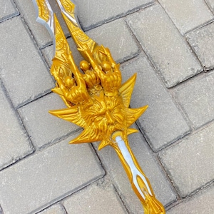 May include: A gold-colored trident with intricate detailing. The trident features a central blade and two side blades, all with a silver accent. The handle is also gold, with a detailed design. The trident is lying on a gray brick surface.