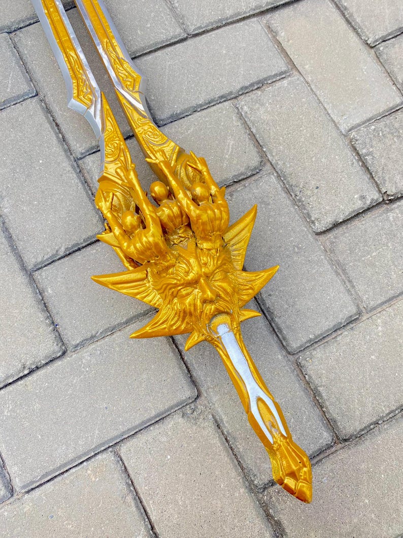 May include: A gold-colored trident with intricate detailing. The trident features a central blade and two side blades, all with a silver accent. The handle is also gold, with a detailed design. The trident is lying on a gray brick surface.