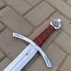May include: A medieval-style sword with a polished steel blade and a brown leather-wrapped handle. The crossguard features the words "CHRISTUS" and "VINCIT". A cross emblem is visible on the blade.