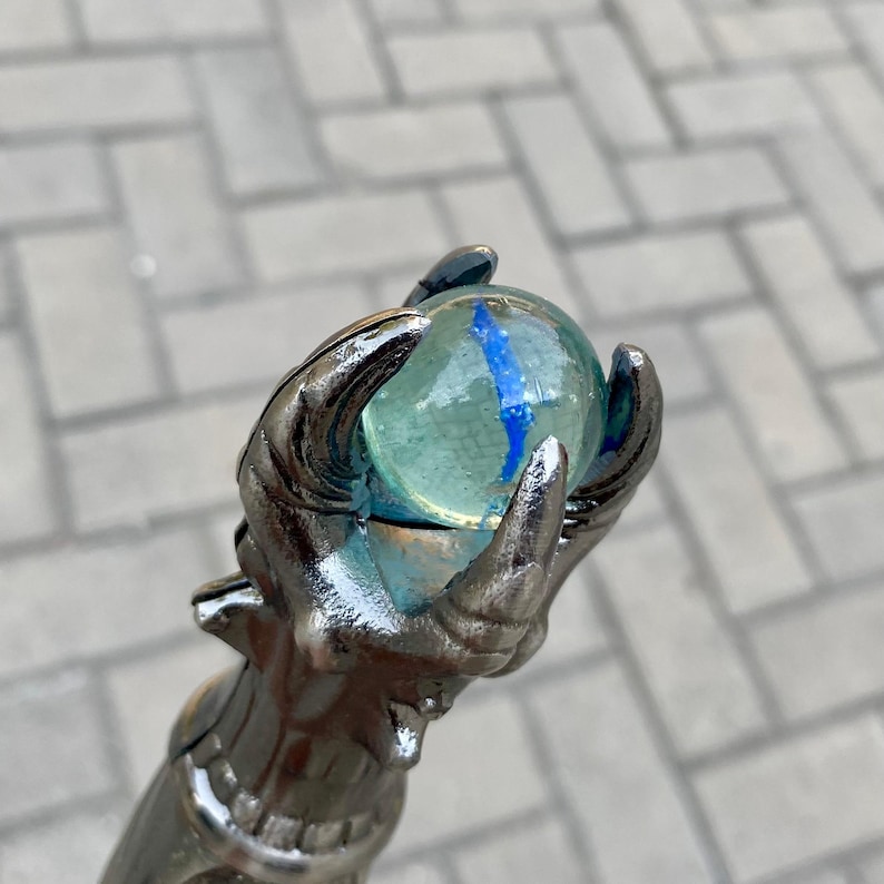 May include: A silver-coloured staff featuring a detailed hand grasping a clear glass orb with blue streaks. The staff has a metallic finish, set against a grey, textured background. The orb is likely a decorative element.