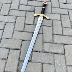 May include: A silver sword with a black leather grip and a gold pommel with a red jewel. The sword is lying on a gray brick surface.