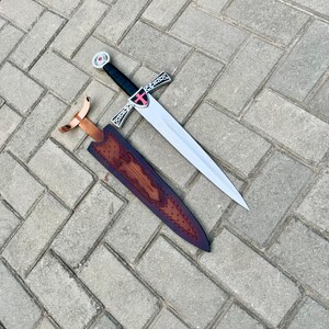 May include: A silver and black sword with a red cross emblem on the hilt, resting beside a brown leather sheath with decorative tooling. The sword has a long, pointed blade and a black handle. The sheath has a copper-colored belt loop.