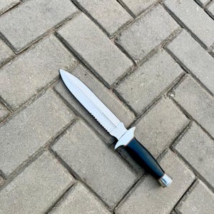 May include: A silver and black dagger with a double-edged blade and a serrated spine. The handle is black with a silver bolster and pommel. The knife is lying on a grey brick surface.