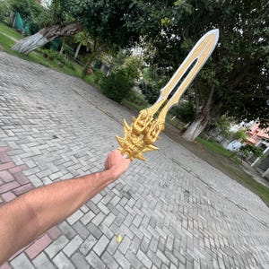 May include: A golden fantasy sword with intricate details, held against a backdrop of greenery and a brick-paved surface. The sword features a detailed hilt and a blade with a contrasting color.
