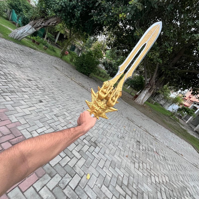 May include: A golden fantasy sword with intricate details, held against a backdrop of greenery and a brick-paved surface. The sword features a detailed hilt and a blade with a contrasting color.