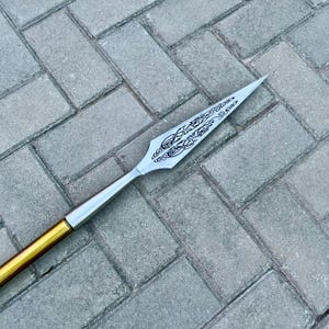 May include: A spearhead with a silver blade and a decorative etched design. The spear has a gold and silver shaft. The spear is lying on a gray brick surface.