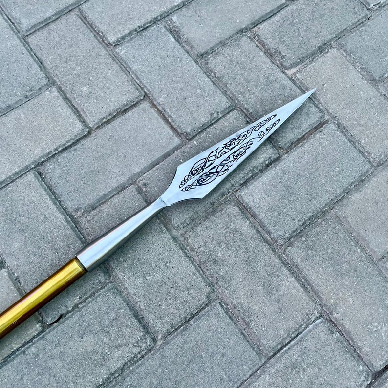 May include: A spearhead with a silver blade and a decorative etched design. The spear has a gold and silver shaft. The spear is lying on a gray brick surface.