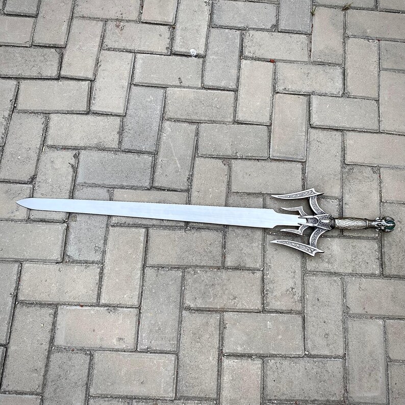 May include: A silver sword with an elaborate handle and guard, resting on a brick surface. The blade is long and straight, with a pointed tip. The handle has detailed carvings and a decorative pommel.