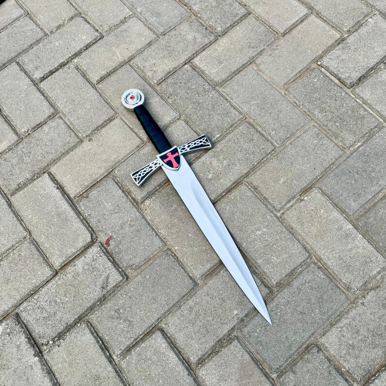 May include: A silver-colored sword with a black handle and a cross-shaped guard. The guard features a red cross emblem. The sword is displayed on a gray brick surface.