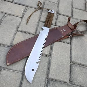 Könnte beinhalten: Eine große, silberne Machete mit gezahnter Kante und braun umwickeltem Griff liegt auf einer braunen Lederscheide. Eine beige Schnur ist am Griff befestigt. Die Machete ist für den robusten Schnitt konzipiert.