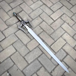 May include: A silver sword with an elaborate hilt and a long, straight blade. The hilt has detailed patterns and a green gem. The sword is resting on a grey brick surface.