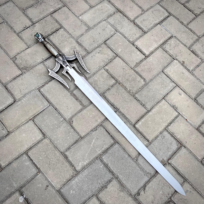 May include: A silver sword with an elaborate hilt and a long, straight blade. The hilt has detailed patterns and a green gem. The sword is resting on a grey brick surface.