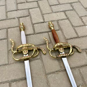 May include: Two ornate swords with gold-colored hilts and silver blades. One sword has a white handle, the other a brown wooden handle. Both feature red accents and intricate designs, suggesting a historical or decorative purpose.