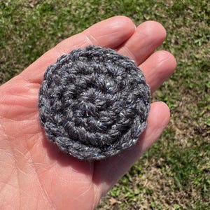 May include: A small, round, crocheted item in shades of gray with silver flecks. The handmade piece is held in a person's hand against a background of green grass. The item's texture is visible, showcasing the yarn's looped construction.