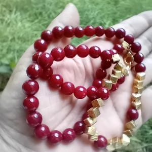 Può includere: Primo piano di due braccialetti. Uno è fatto di perline rotonde rosso scuro. L'altro braccialetto ha perline dorate a forma di cubo. I braccialetti sono tenuti in una mano su uno sfondo verde sfocato.
