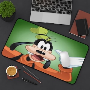 Puede incluir: Alfombrilla de ratón rectangular con una imagen de dibujos animados de Goofy con su atuendo clásico naranja y verde, señalando con una mano enguantada de blanco. El fondo es un degradado de verde. Un ratón de ordenador descansa sobre la alfombrilla.