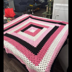 May include: A square crochet blanket featuring a geometric pattern of concentric diamonds. The design consists of alternating bands of light pink, white, dark pink, and black yarn, worked in a traditional granny square stitch construction.