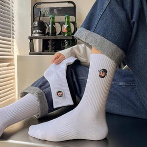 May include: White crew socks with a cartoon face embroidered on each sock. The socks are folded and displayed on a person's legs.