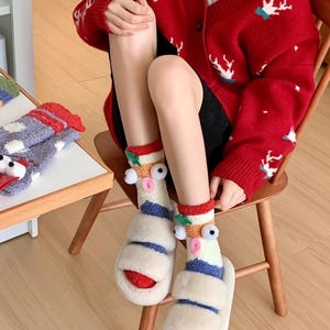 Può includere: Calzini originali con occhi mobili e dettagli a righe, abbinati a sandali bianchi e soffici con suole rosse. La persona indossa anche un cardigan rosso con disegni di cervi bianchi.