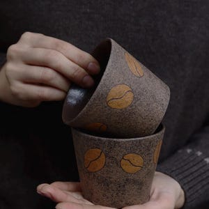 May include: Two brown ceramic coffee cups with a speckled texture and coffee bean designs. The cups are stacked, with one slightly tilted, revealing a dark interior. The cups are held in a person's hands, suggesting a cozy, warm beverage experience.