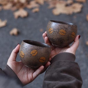 May include: Two small, round, brown ceramic cups with a speckled texture. Each cup features three coffee bean designs in a lighter brown color. The cups are held in hands, with a blurred background of fallen leaves.