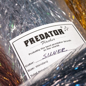 Op de afbeelding: Een verpakking zilverkleurig Predator Flasher synthetisch vliegbindmateriaal met een wit label met de tekst PREDATOR Flasher, Probably the best predator brush in the world, Color: SILVER, Size: standard. Het materiaal is dun en reflecterend.