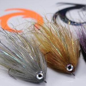 Pode incluir: Três iscas de pesca com fibras de pelo sintético em cinza, castanho e preto com fios de ouropel iridescente. Cada isca tem um olho prateado e preto, um anzol de metal afiado e está posicionada à frente de caudas de plástico laranja e preto.