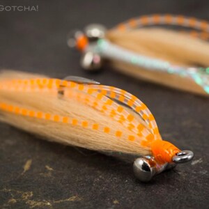 Pode incluir: Dois iscas de pesca com mosca laranja e branca com pernas de silicone e uma corrente de contas. O texto "Sili Legs Gotcha!" é visível na imagem.