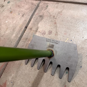 May include: A green metal rake with a metal handle. The rake has seven tines and is made in California. The text "Alchemy Trail Tools Made in California" is printed on the rake.