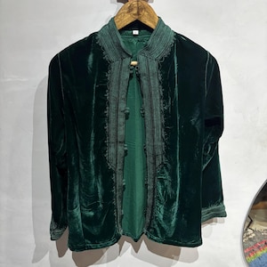 May include: A dark green velvet jacket with detailed embroidery on the collar and front. The jacket, with long sleeves, hangs on a wooden hanger. The design includes a high collar and decorative trim, showcasing a rich, textured appearance.