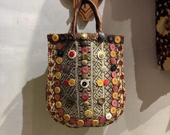 Puede incluir: Un pequeño bolso de mano marrón con un diseño único. El bolso está cubierto de botones coloridos de varios tamaños y formas. El bolso tiene un asa de cuero marrón.