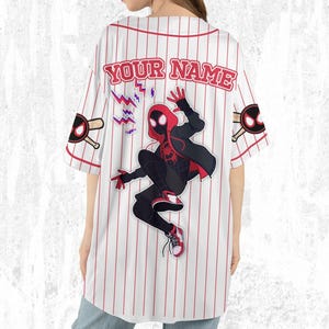 May include: White baseball jersey with red pinstripes and the text "YOUR NAME" across the back. Features a graphic of a superhero in a black and red suit. Includes baseball bat and ball graphics on the sleeves.