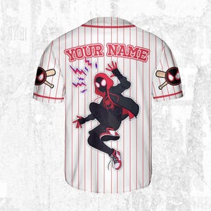 May include: White baseball jersey with red pinstripes, featuring a graphic of a superhero in a dynamic pose. The jersey includes the text "YOUR NAME" across the back, with superhero-themed logos on the sleeves.